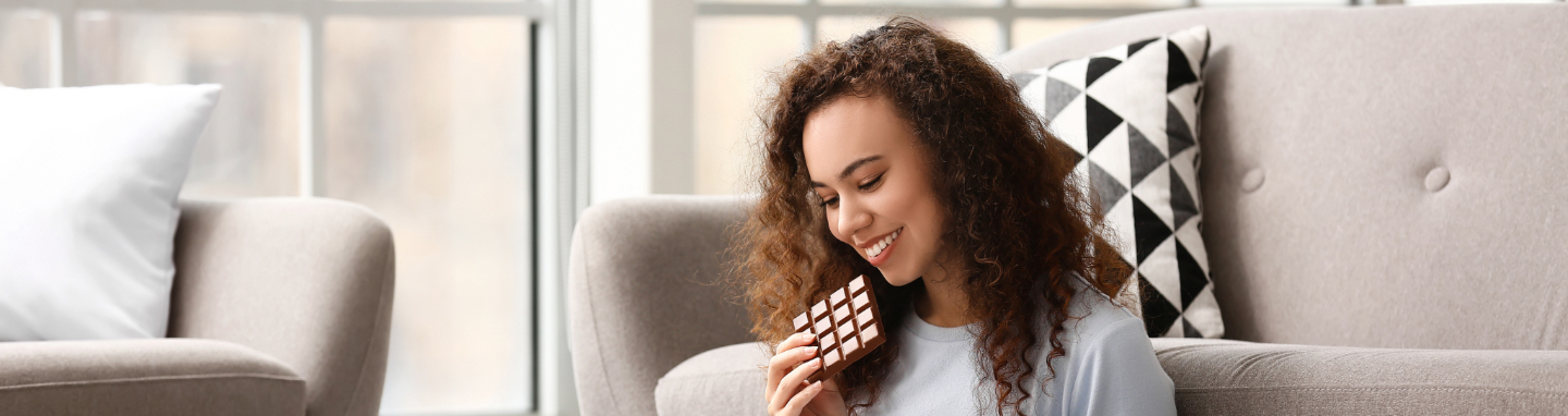 Woman eating a chocolate bar while sitting on the carpet an working from her laptop.