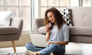 Woman eating a chocolate bar while sitting on the carpet an working from her laptop.